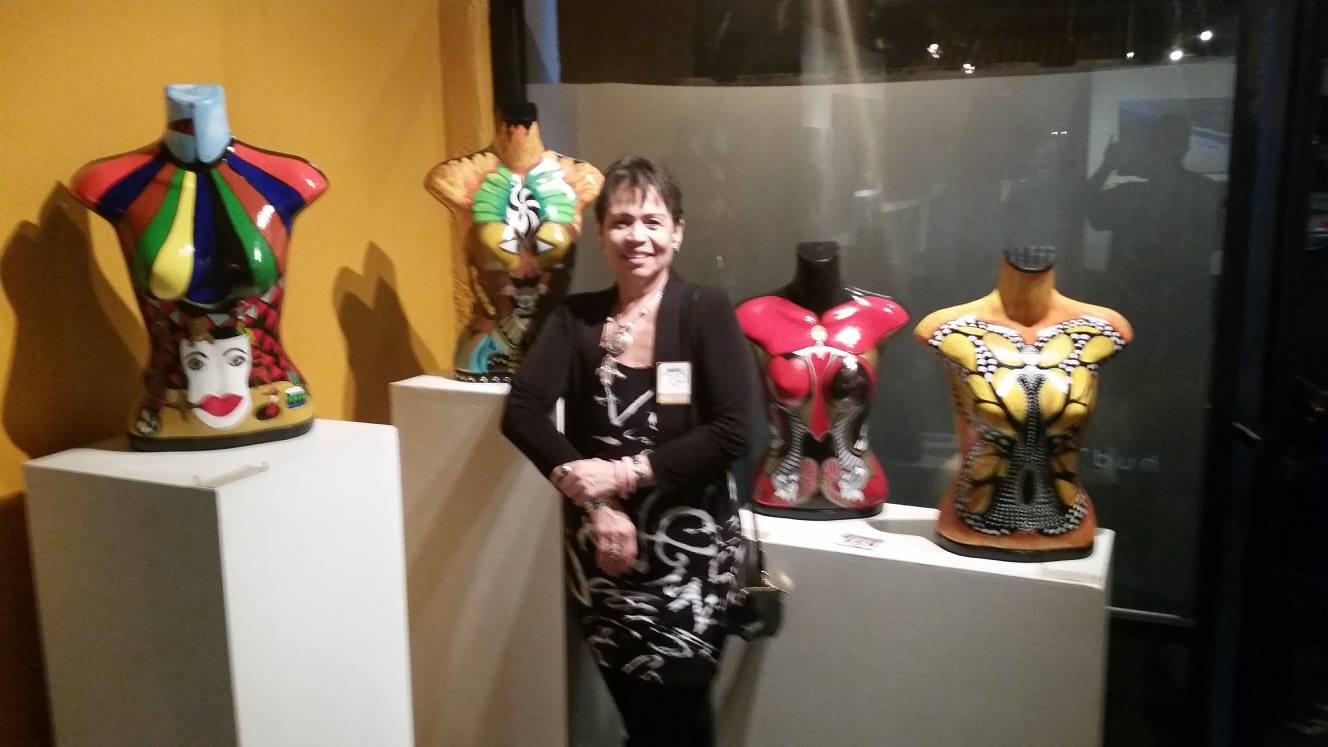 Woman standing in front of colorful, decorative sculptures on display, with a dark background and spotlights highlighting the art pieces.