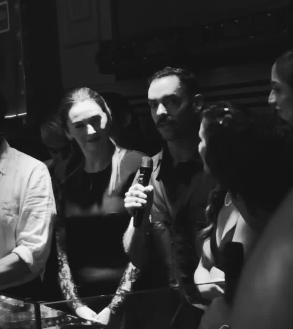 Group of people, one holding a microphone, in a dark indoor setting, some smiling, some looking serious.