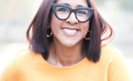 Smiling woman with shoulder-length dark hair, wearing large glasses, gold hoop earrings, and a yellow top outdoors.