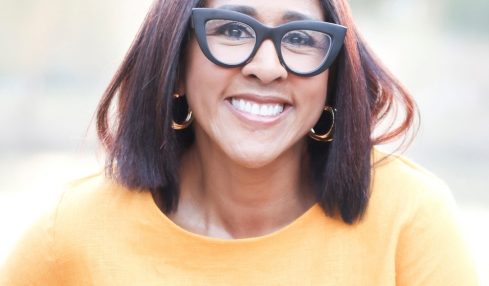 Smiling woman with shoulder-length dark hair, wearing large glasses, gold hoop earrings, and a yellow top outdoors.
