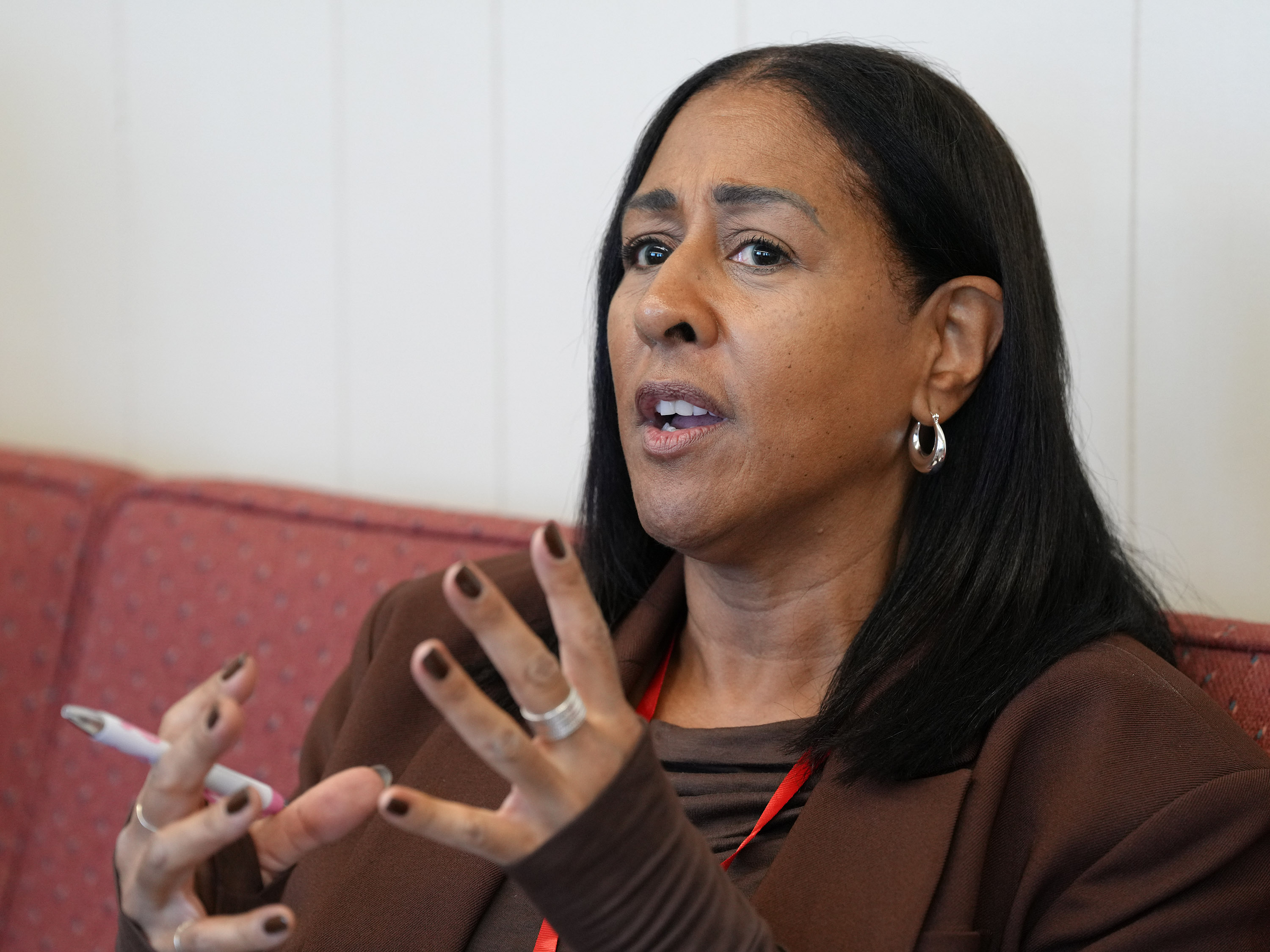 Woman with black hair and hoop earrings gestures with her hands while speaking, seated on a red couch against a white wall.