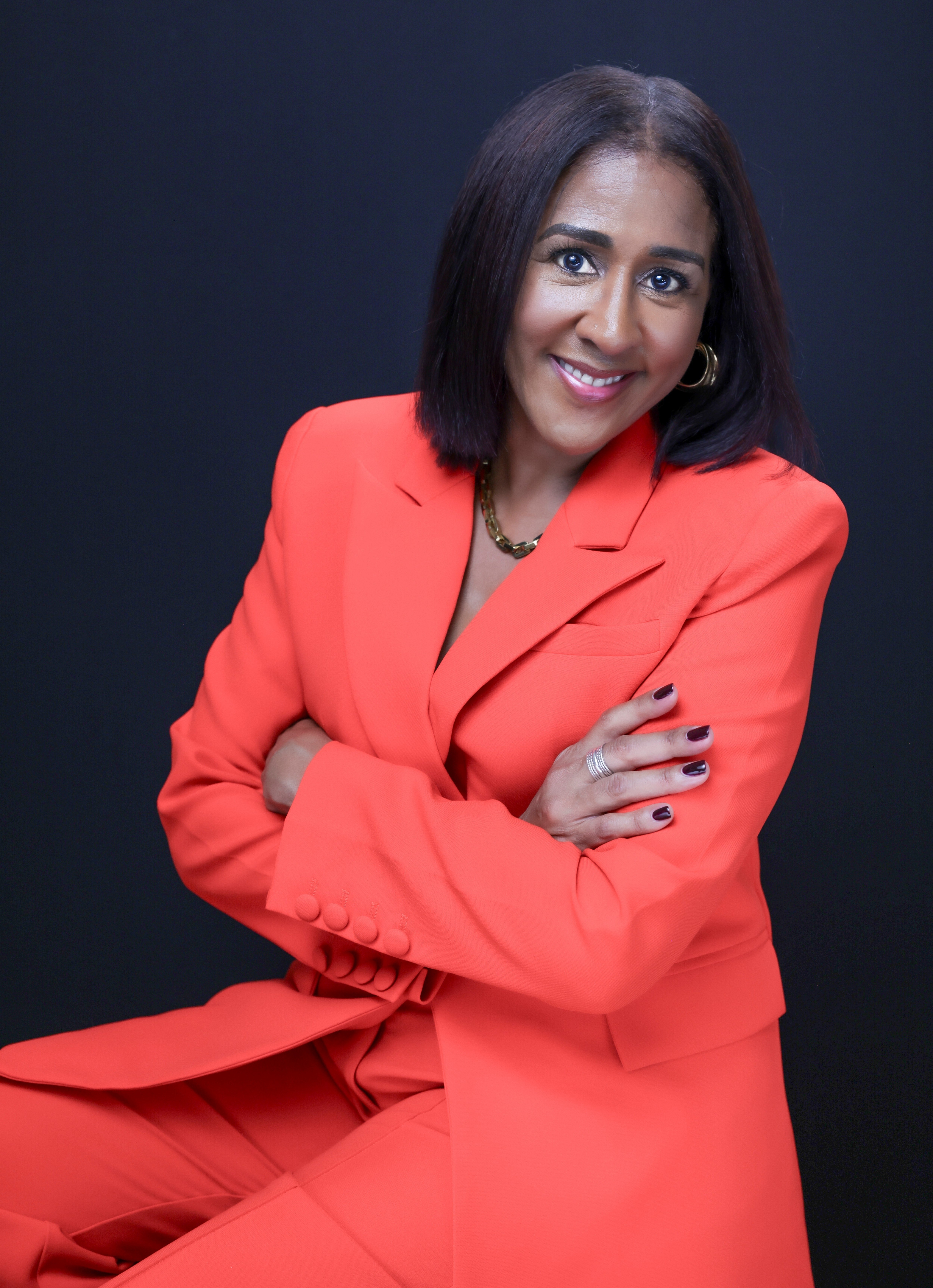 Woman with shoulder-length dark hair smiling, wearing a red blazer and matching pants, crossed arms, dark background.