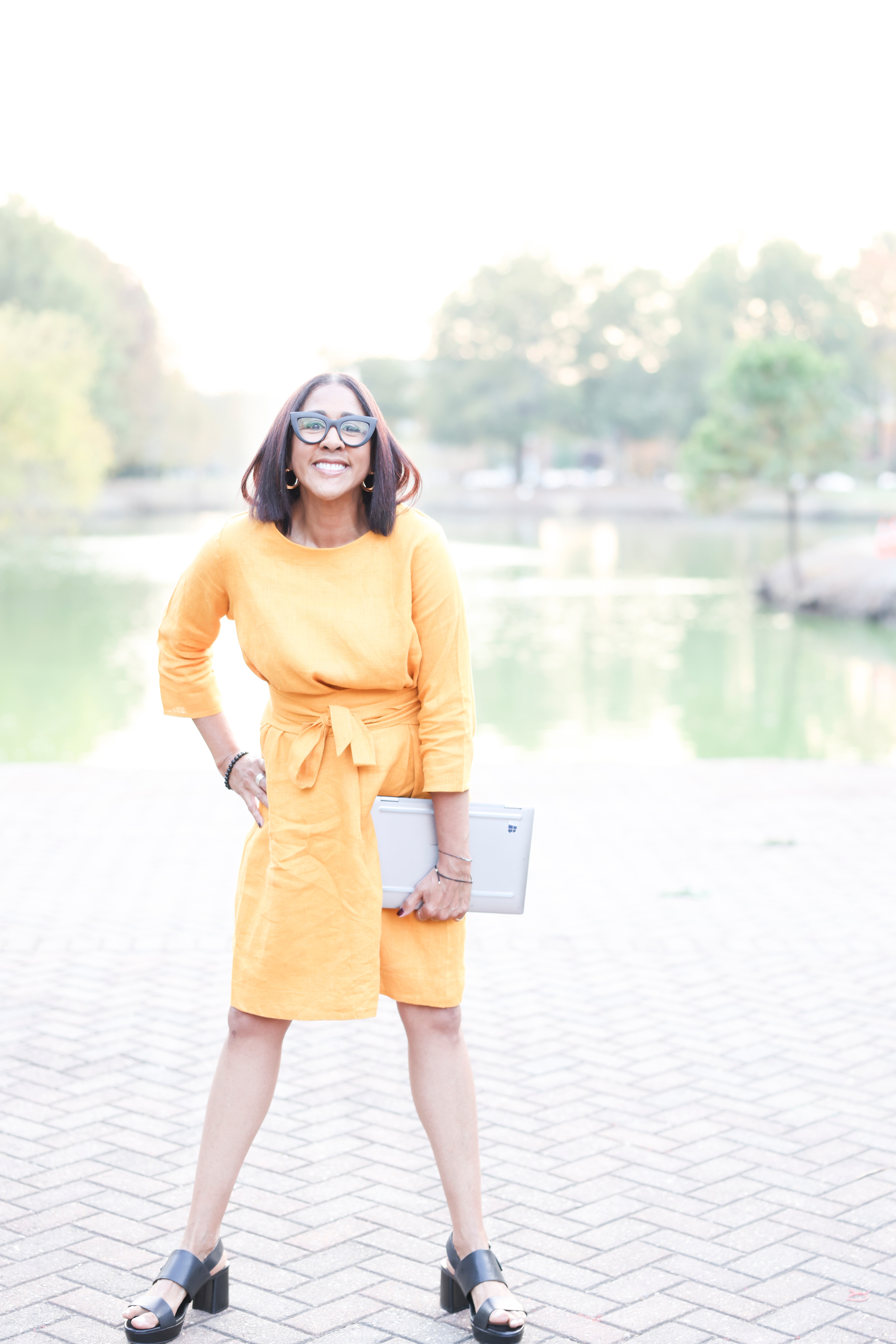 Woman in yellow dress holding a laptop, standing outdoors near water, smiling, wearing sunglasses, with trees in background.
