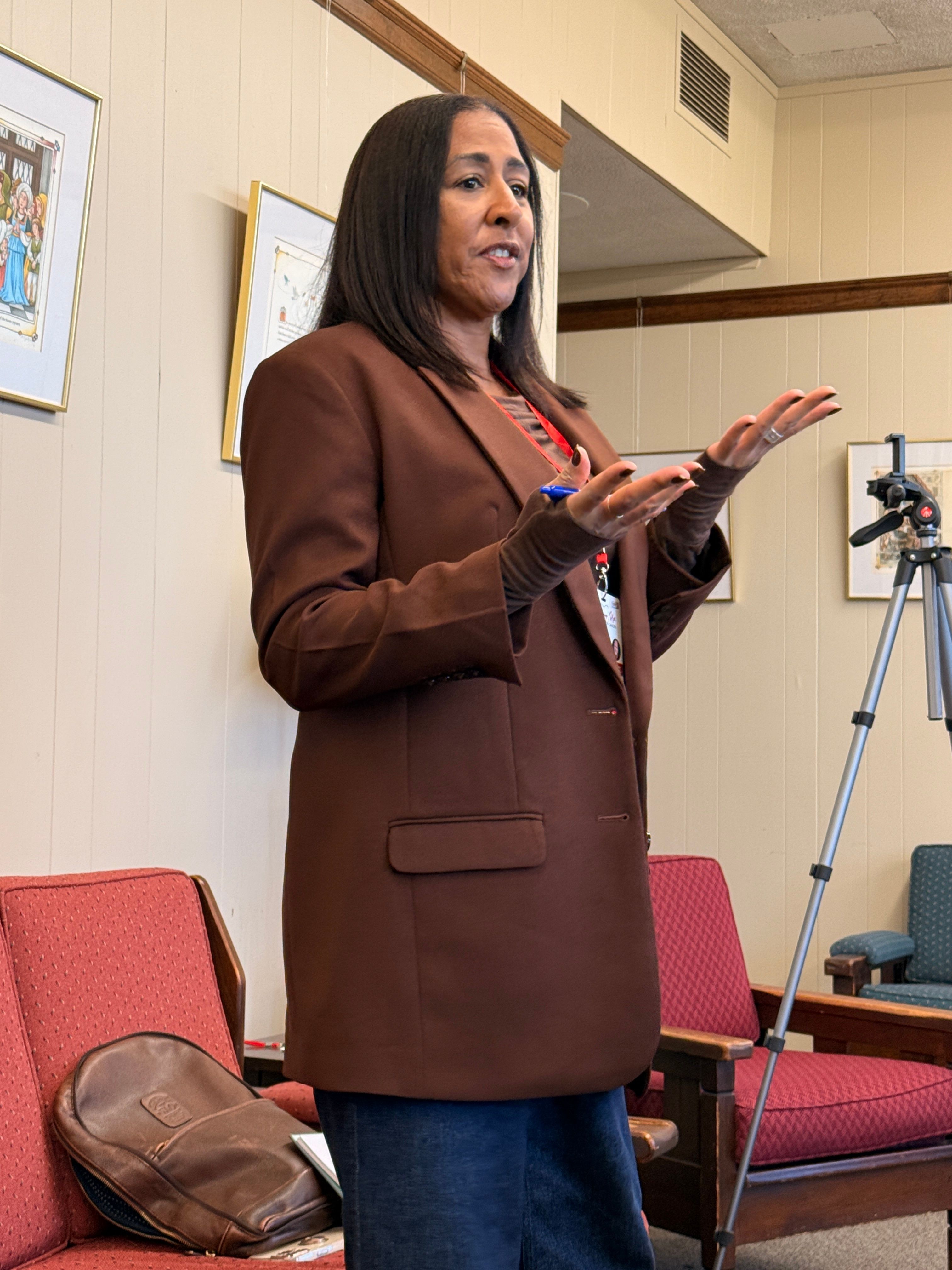 Woman standing in a room, gesturing with hands, wearing a brown coat and blue jeans, with chairs and a tripod in the background.