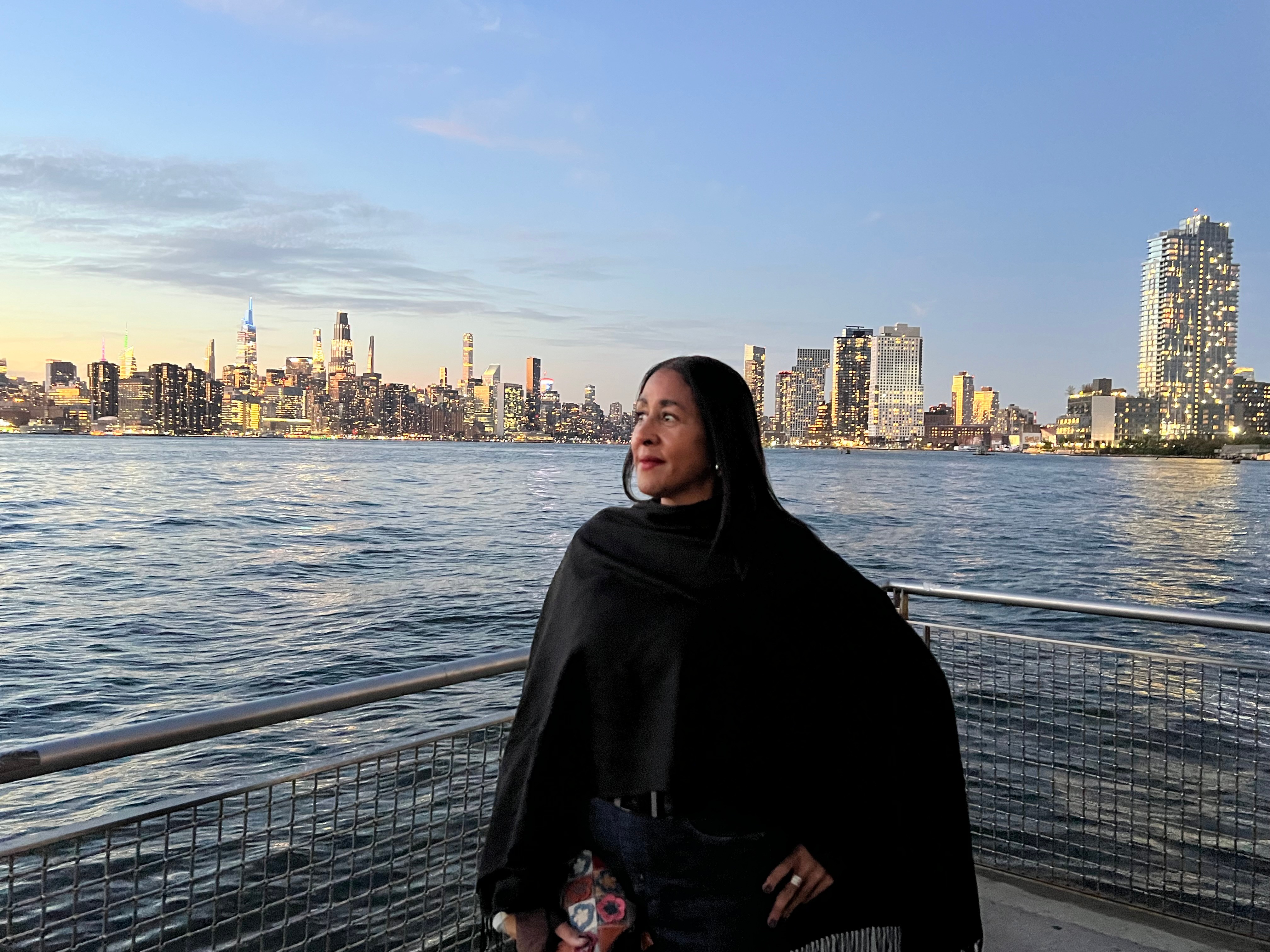 Woman standing by a waterfront railing with city skyline in background, smiling and looking to the side.