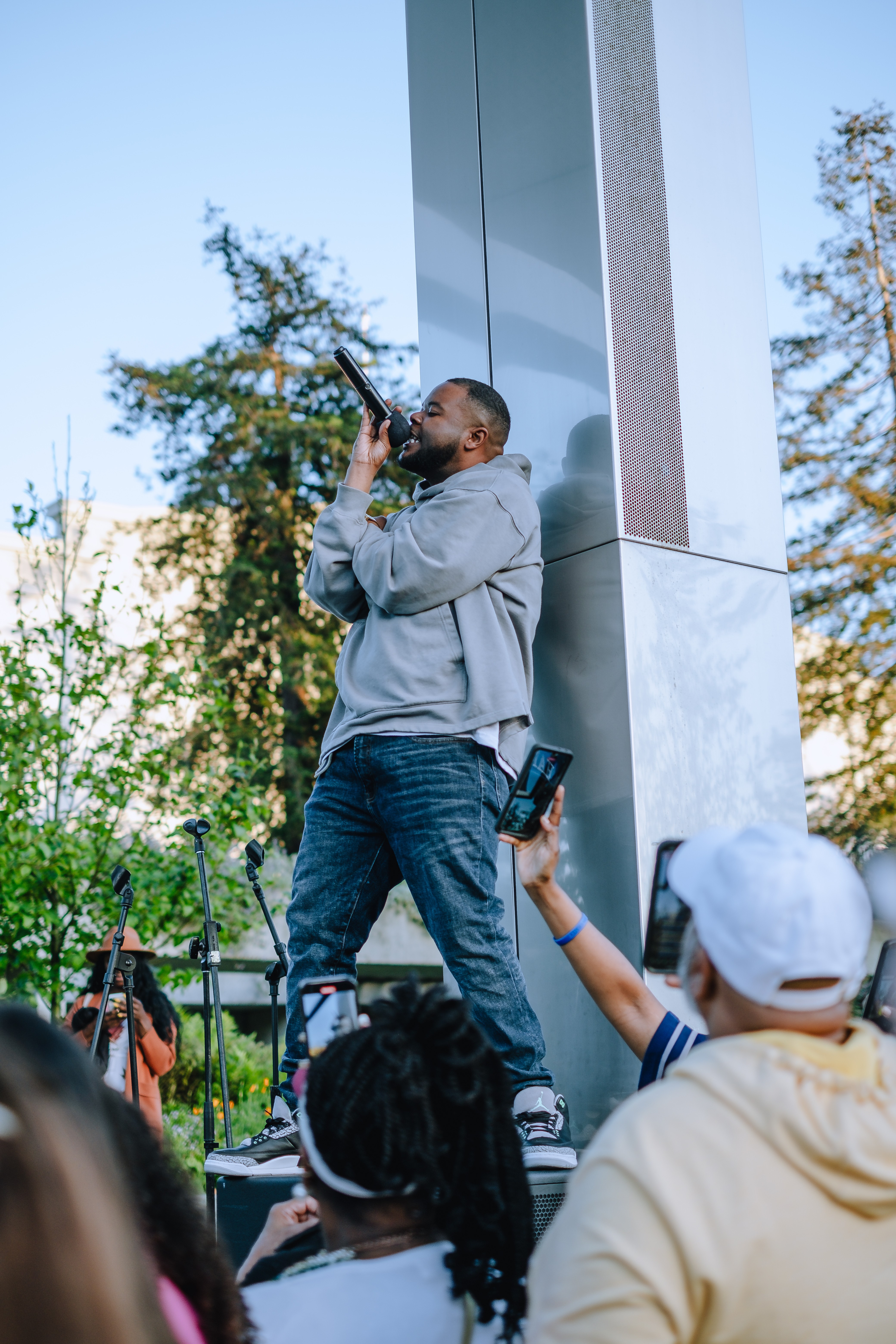 Man speaking into microphone on outdoor stage with audience, trees, and a building in background.