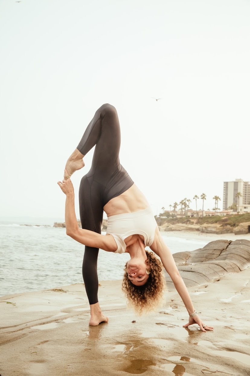 Person performing a yoga pose on the beach, with buildings and palm trees in the background.