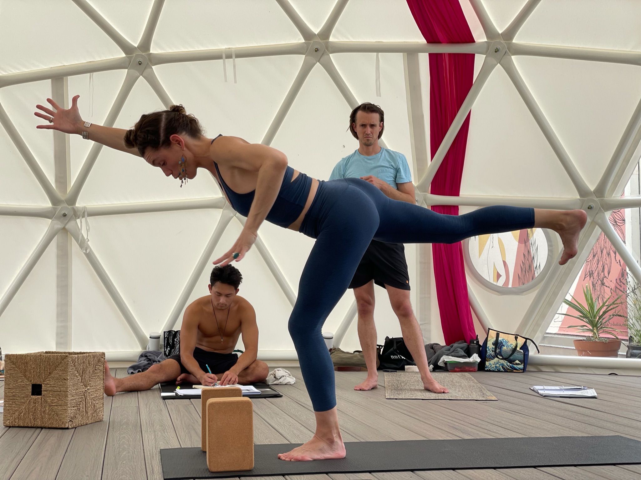 Woman in yoga pose balancing on one leg inside a geodesic dome, with two men nearby, one sitting and one standing.