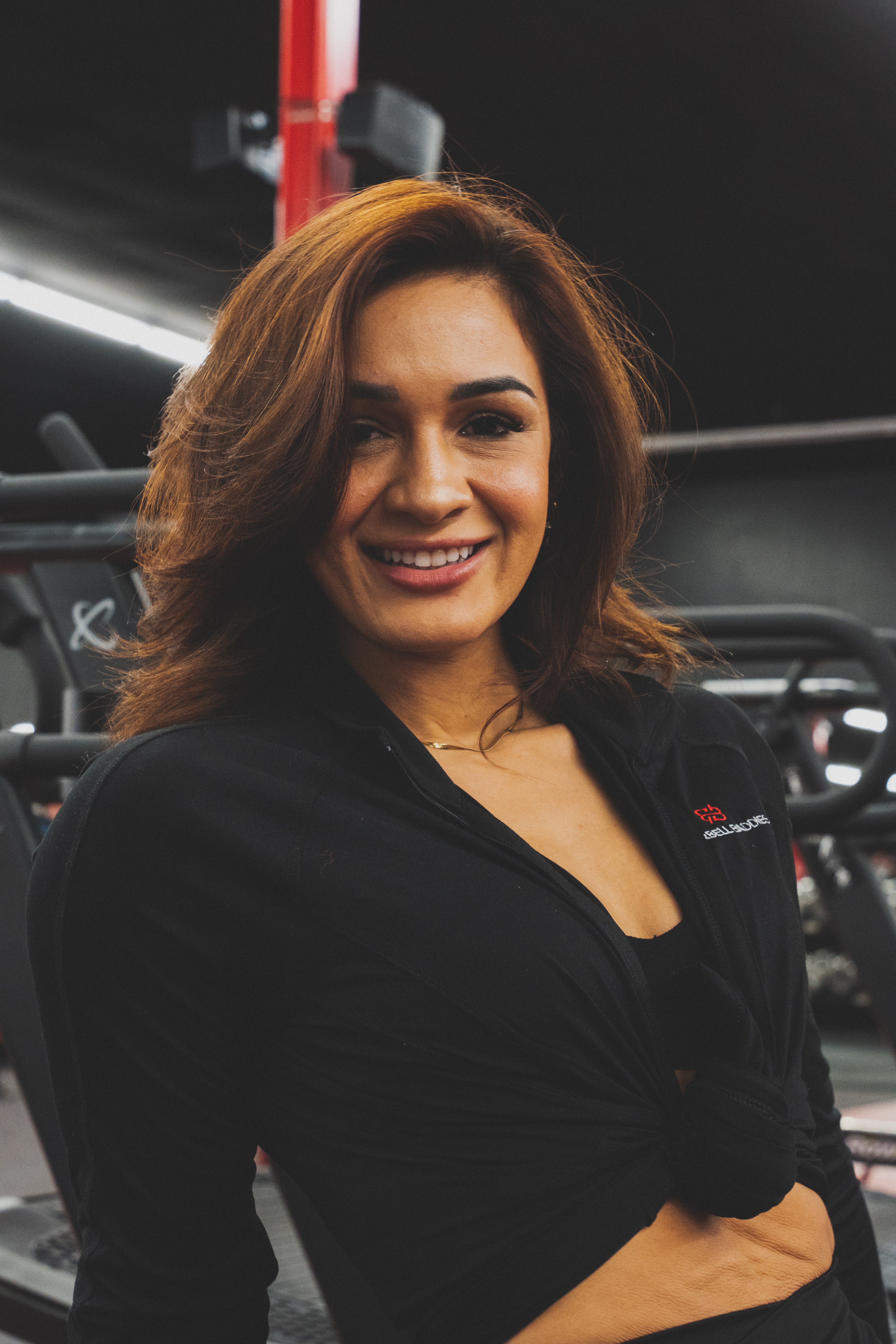 Woman with shoulder-length hair smiling in a gym, wearing a black top, with gym equipment in background.