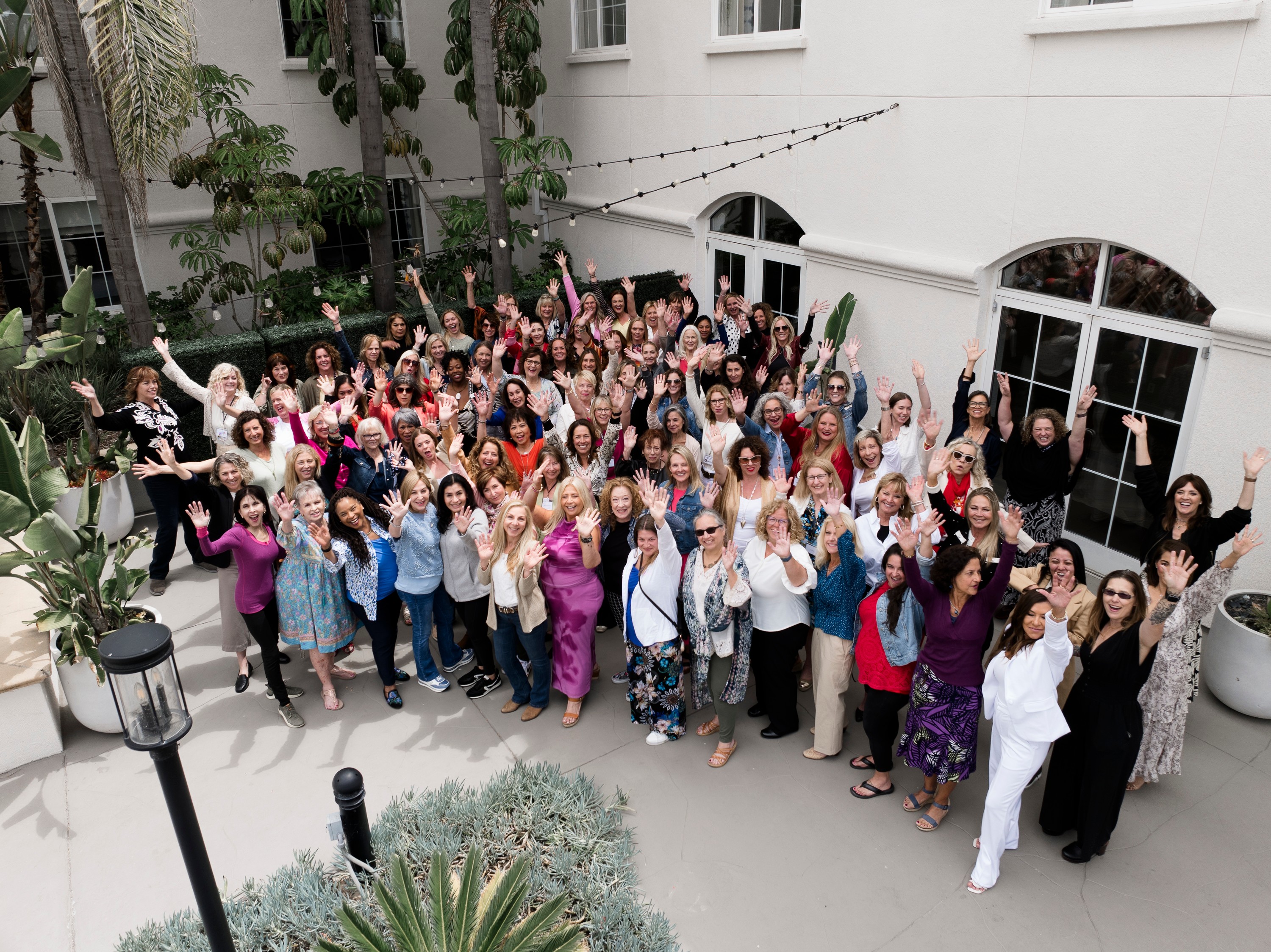 Large group of people gathered outdoors in a courtyard, smiling and waving at the camera.