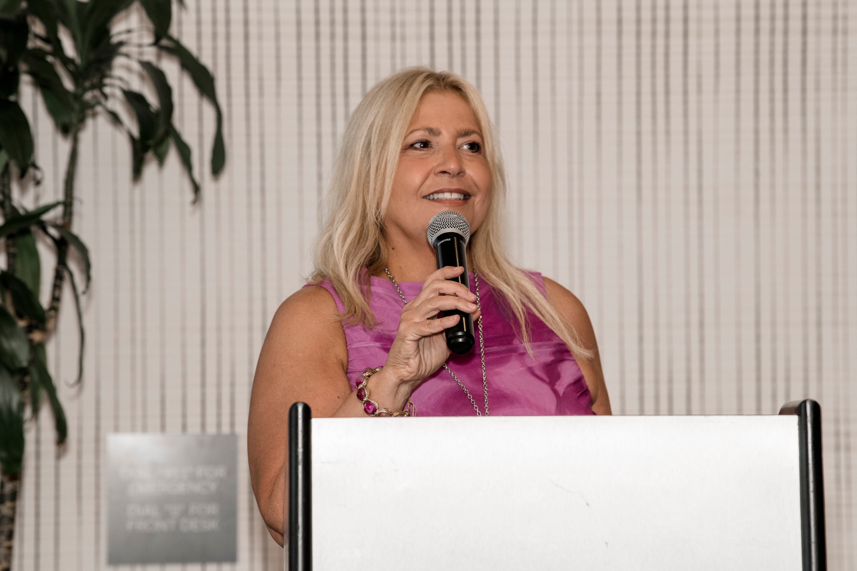 Woman with long blonde hair speaking into a microphone, wearing a pink sleeveless top, smiling, standing behind a white podium.