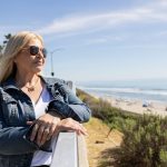 Woman with blonde hair wearing sunglasses and a denim jacket stands outdoors near the beach, with ocean and sky in background.