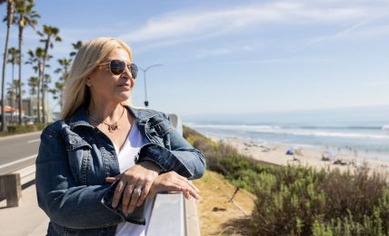 Woman with blonde hair wearing sunglasses and a denim jacket stands outdoors near the beach, with ocean and sky in background.