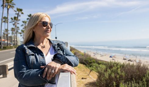 Woman with blonde hair wearing sunglasses and a denim jacket stands outdoors near the beach, with ocean and sky in background.