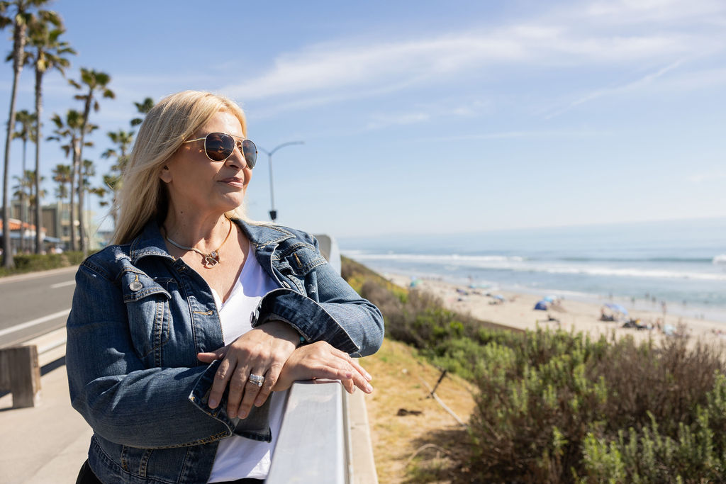 Woman with blonde hair wearing sunglasses and a denim jacket stands outdoors near the beach, with ocean and sky in background.