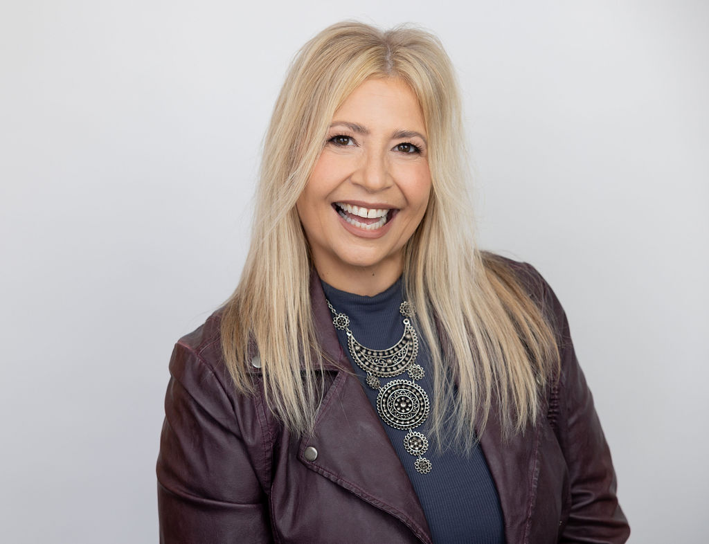Woman with blonde hair smiling, wearing a dark top and a leather jacket, against a plain light background.