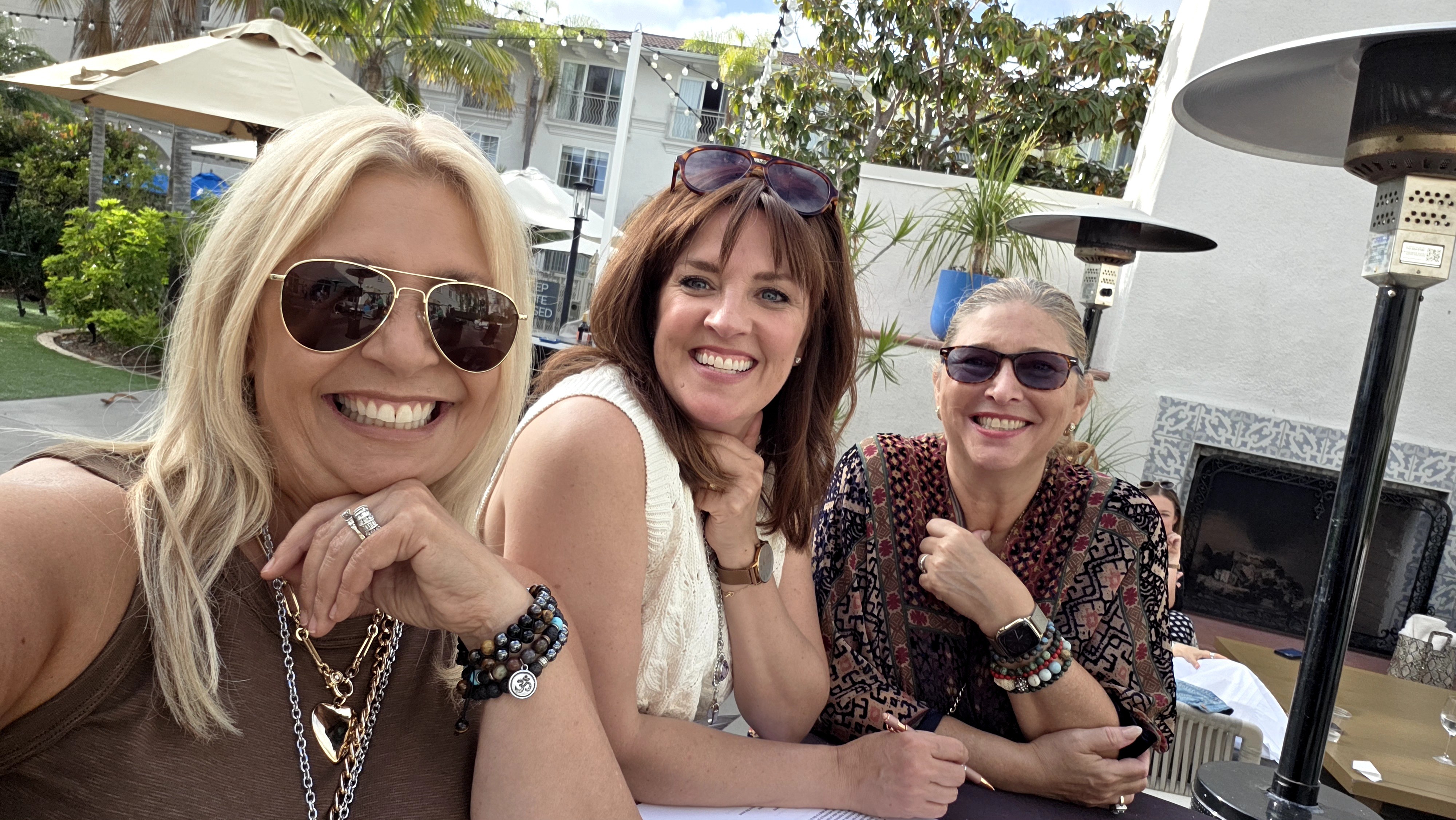 Three women smiling outdoors, wearing sunglasses, sitting at a table with outdoor heaters, trees, and buildings in background.