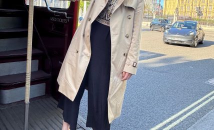 Woman standing on a city street near a bus stop, with buildings and a blue sky in the background.