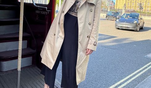 Woman standing on a city street near a bus stop, with buildings and a blue sky in the background.