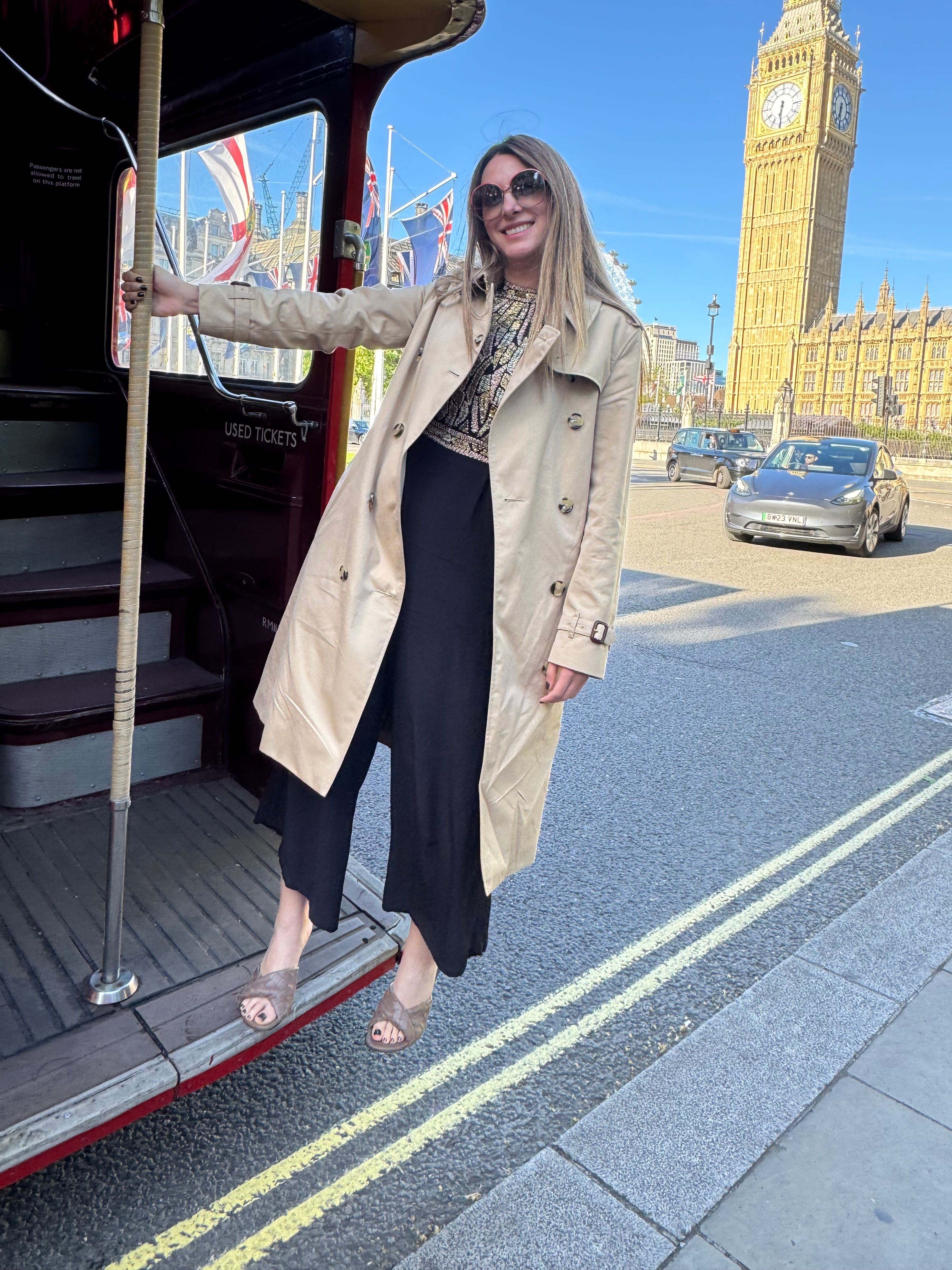 Woman standing on a city street near a bus stop, with buildings and a blue sky in the background.