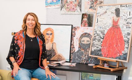 Woman sitting on a stool smiling, surrounded by colorful paintings and artwork displayed on the wall and table.