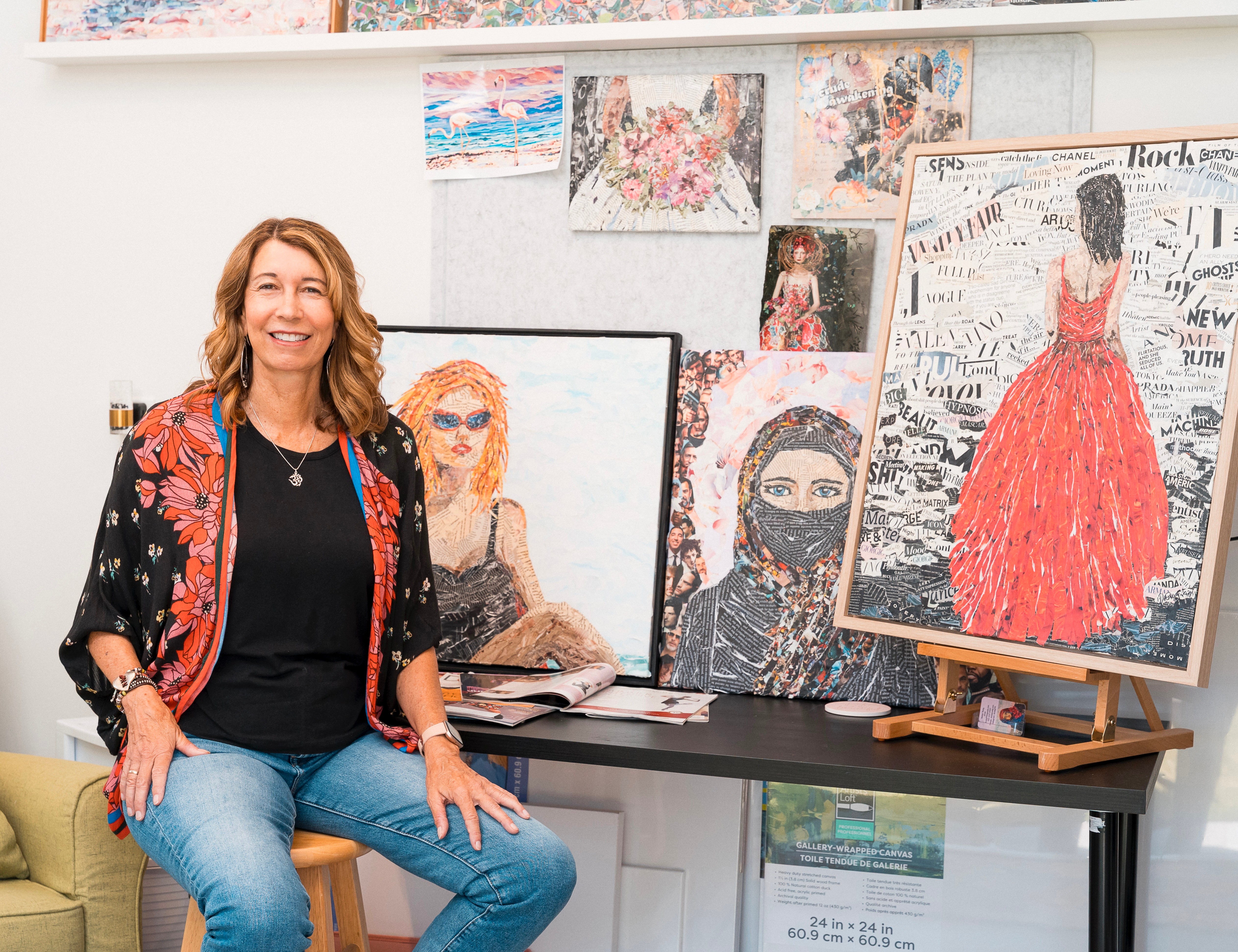 Woman sitting on a stool smiling, surrounded by colorful paintings and artwork displayed on the wall and table.