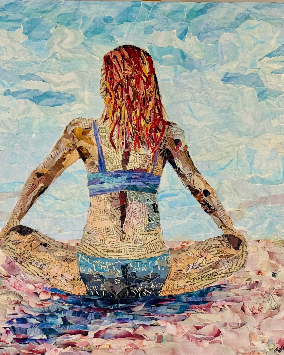 Person sitting cross-legged on a beach, facing away, with red hair, wearing a blue top and denim shorts, ocean background.