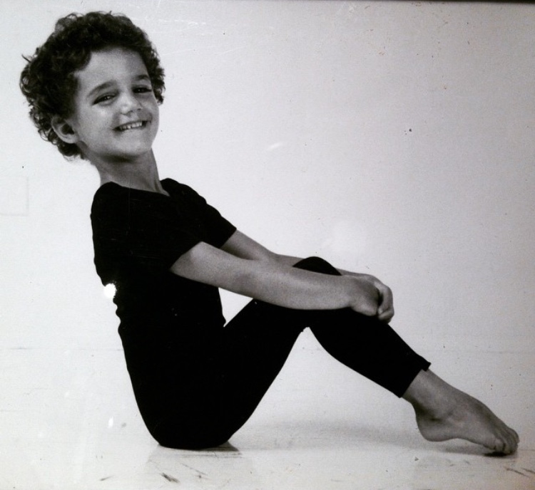 Child with curly hair smiling, sitting with legs extended, wearing a black outfit, against a plain background.