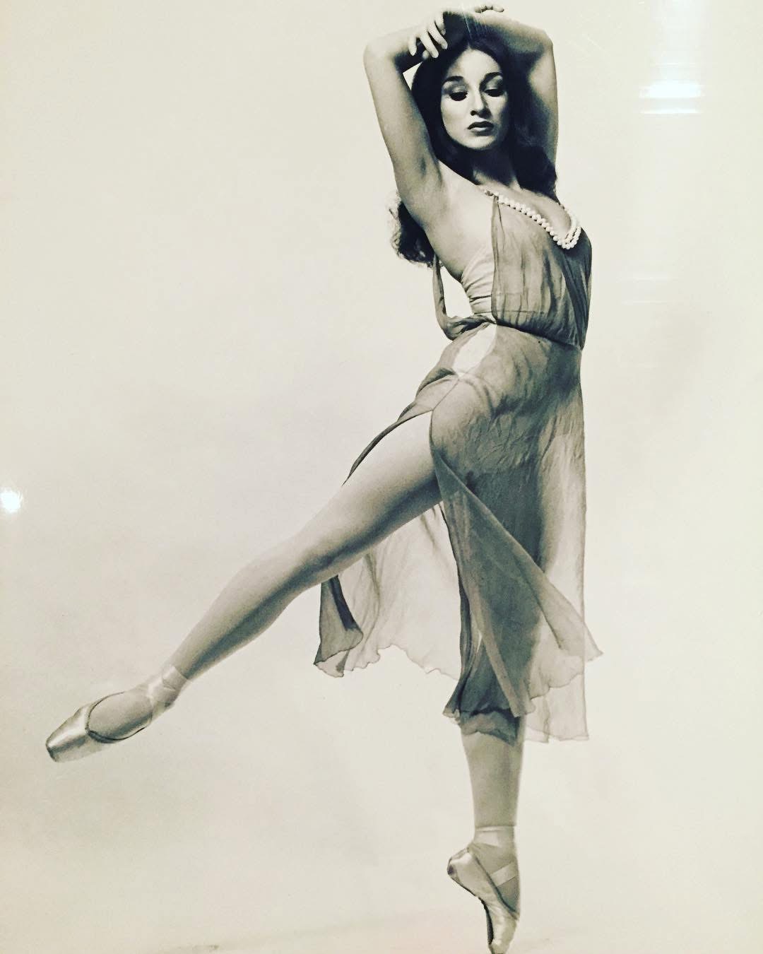 Ballet dancer in a flowing dress poses on pointe with one leg extended and arms raised above her head.