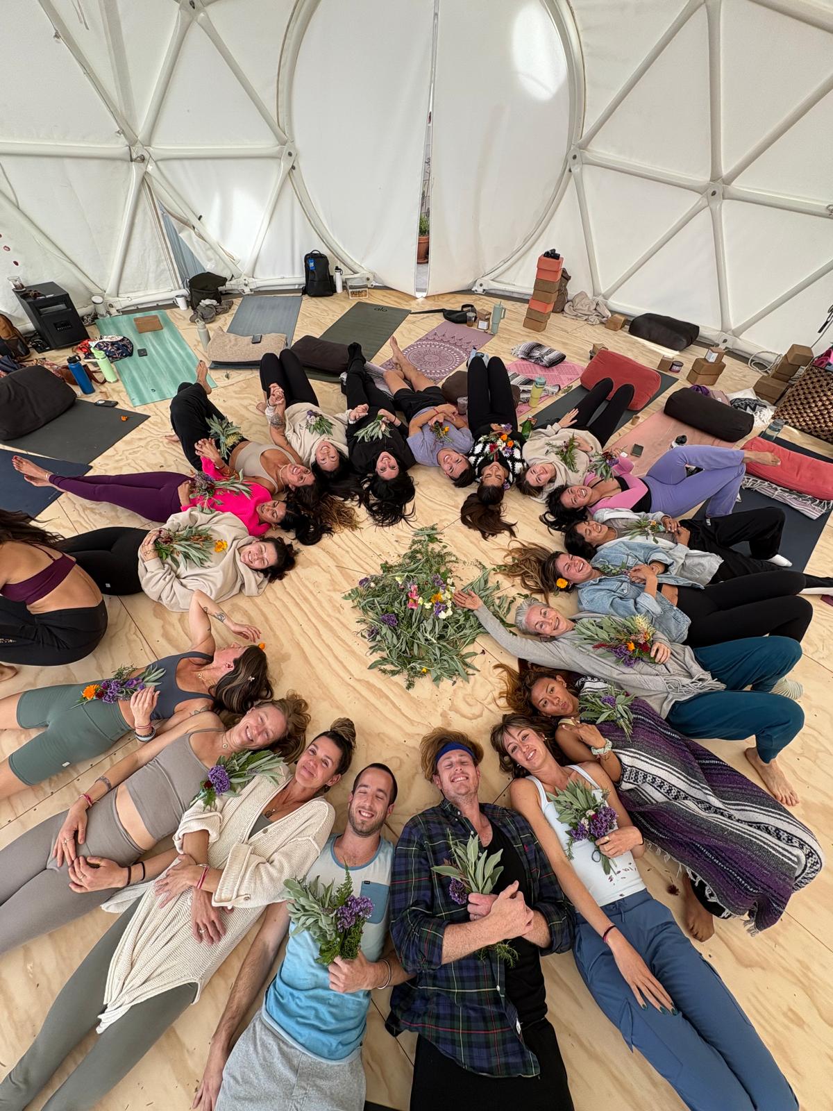 Group of people lying in a circle on a wooden floor inside a tent, with a floral centerpiece in the middle, smiling and relaxing.