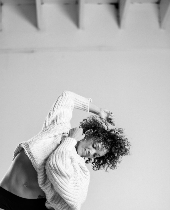 Person with curly hair wearing a sweater, head tilted, arm raised, in a room with ceiling beams