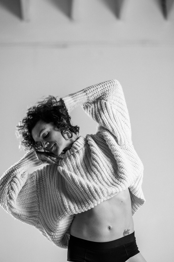 Person with curly hair wearing a large sweater, lifting it to reveal their midriff, smiling, in black and white.