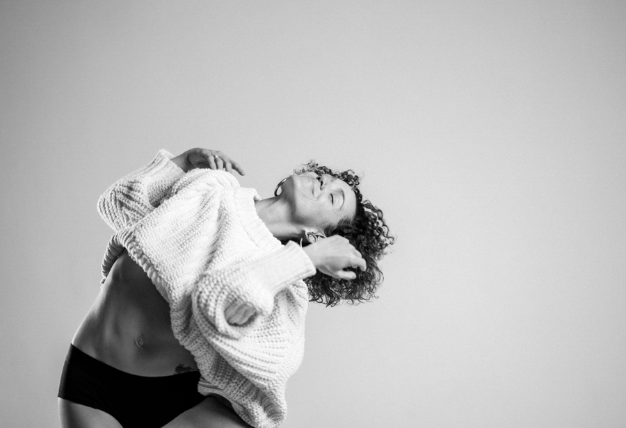Person with curly hair wearing a sweater, posing with eyes closed against a plain background.
