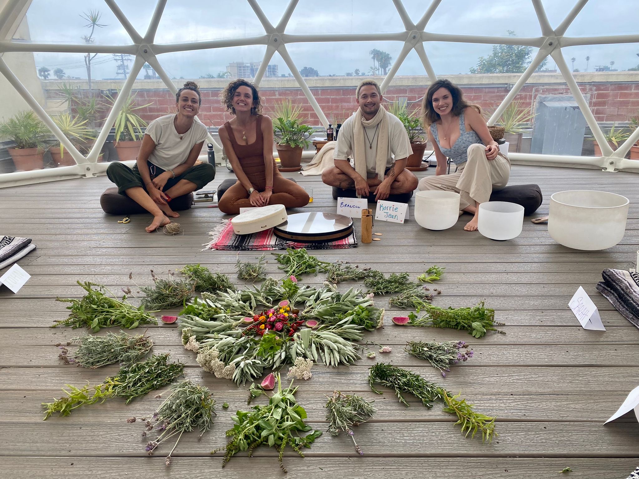 Four people sit on a wooden floor inside a geodesic dome, with musical instruments and plants arranged in front of them.