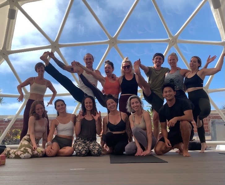 Group of 13 people posing indoors with a geometric glass ceiling, some standing, some sitting, smiling and making poses.