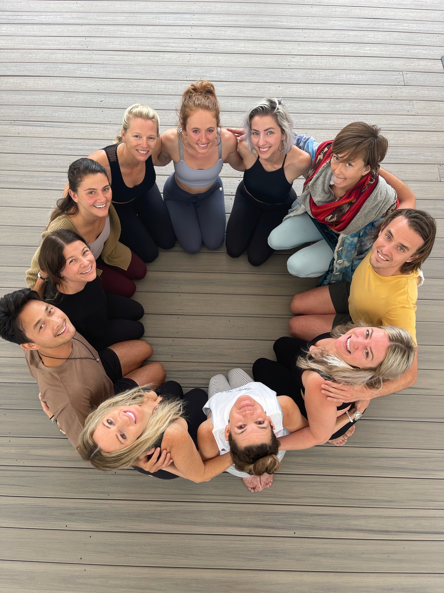Group of ten women sitting in a circle on wooden floor, looking up at camera, smiling.