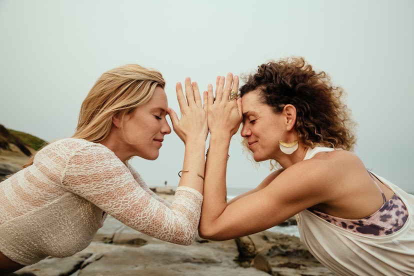 Two women face each other with hands touching, eyes closed, outdoors near a rocky shoreline, overcast sky.