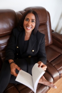 Woman with dark hair smiling, sitting on a brown leather couch, holding an open book, wearing a black blazer.