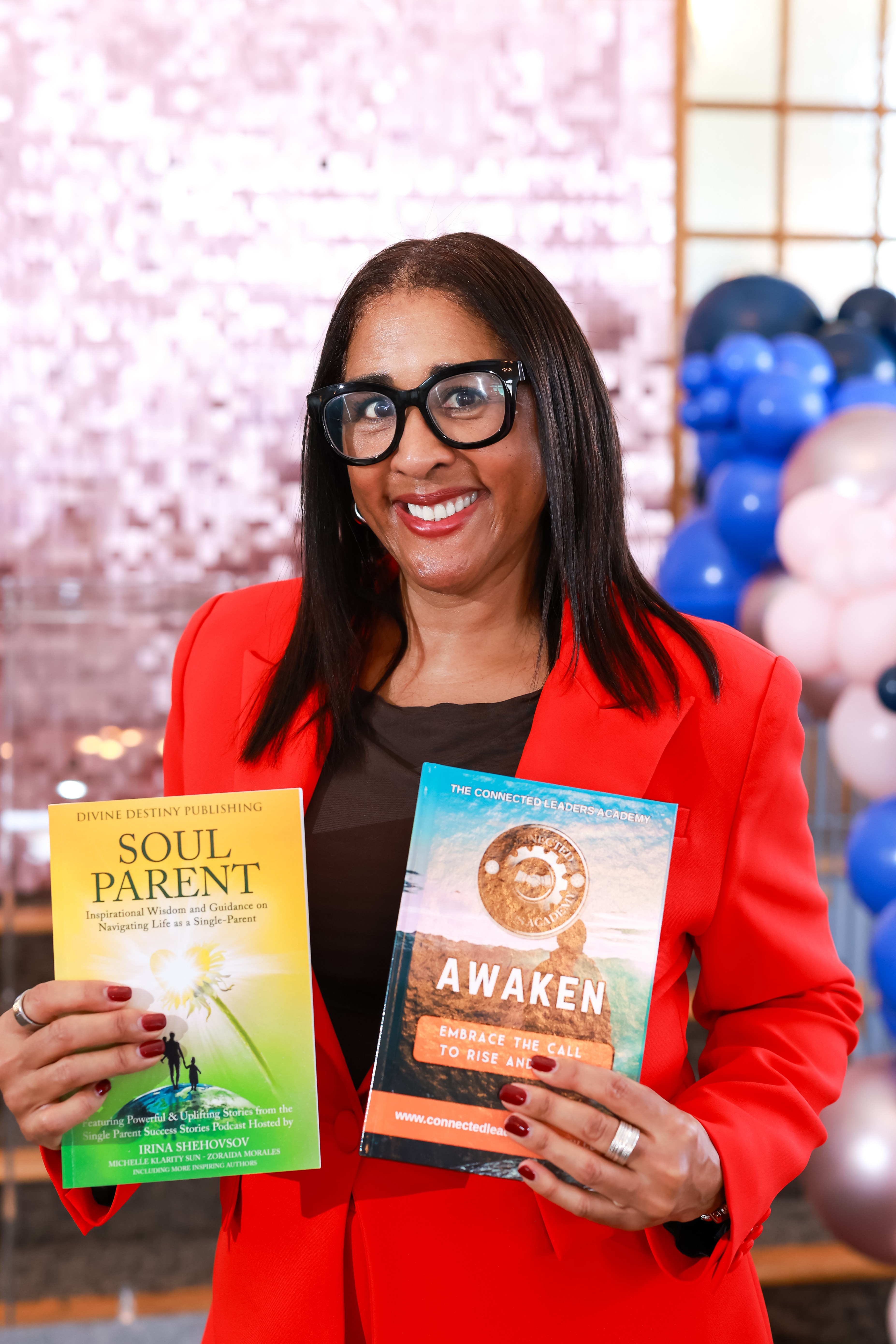 Woman with glasses in red blazer holding two books, smiling, indoor setting with balloons in background.