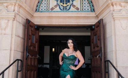 Woman in a green gown with a high slit standing in front of an ornate building entrance, hands on hips.