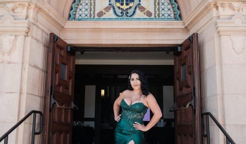 Woman in a green gown with a high slit standing in front of an ornate building entrance, hands on hips.