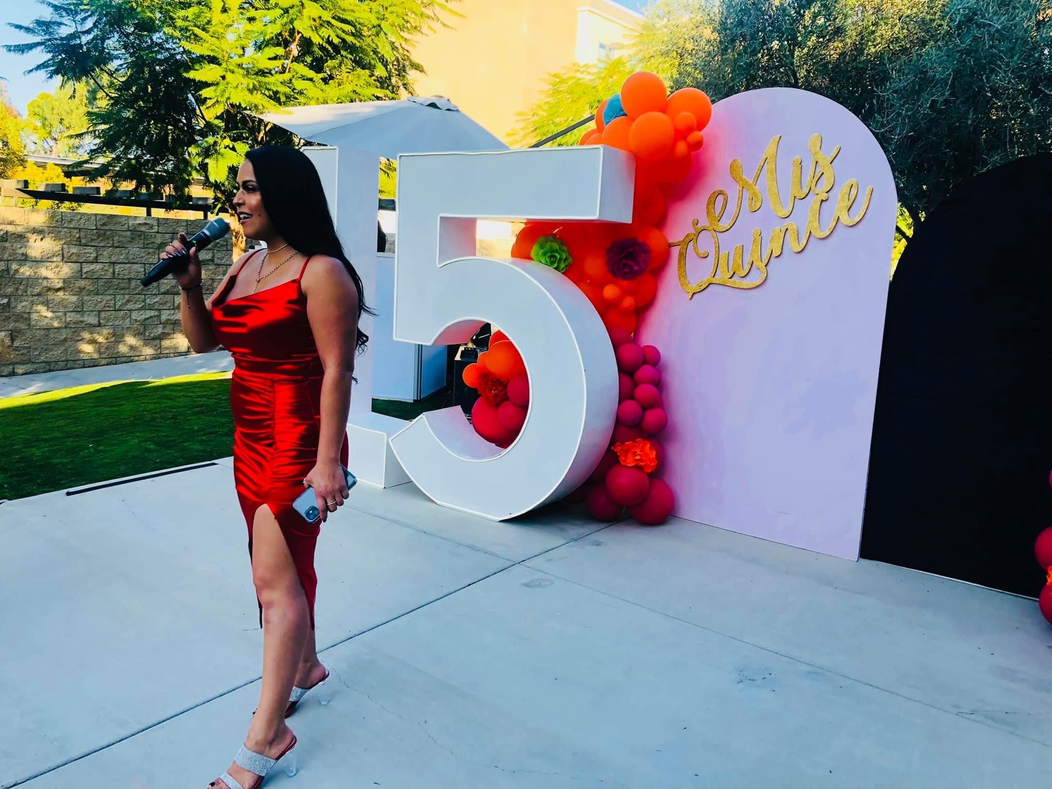 Woman in red dress holding microphone at celebration event with large number five and colorful decorations behind her.