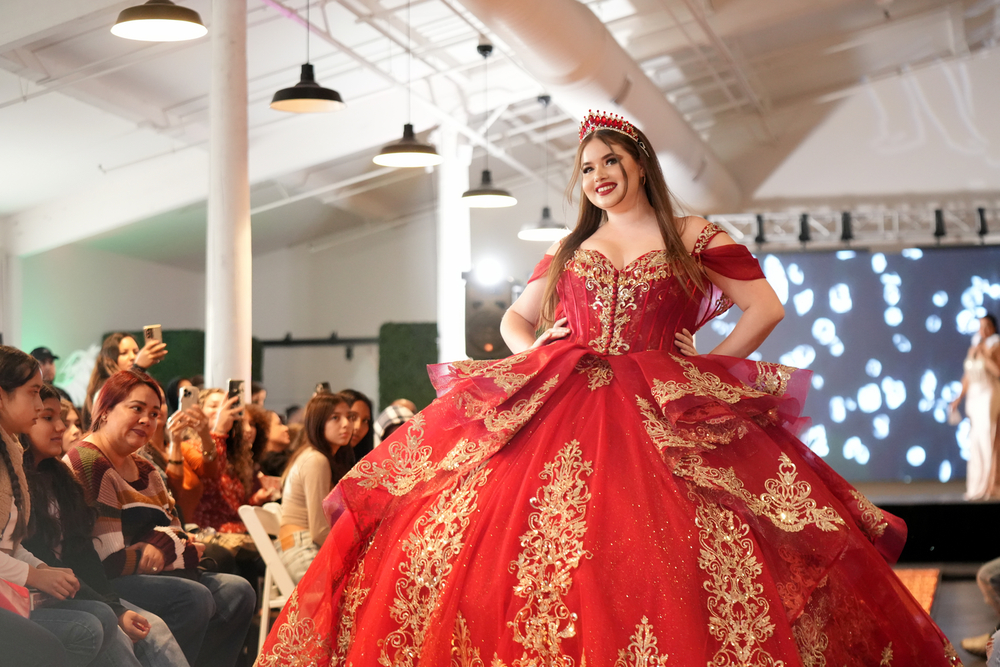 A woman in a red and gold gown with a crown, smiling on stage at a fashion show or event, audience in background.