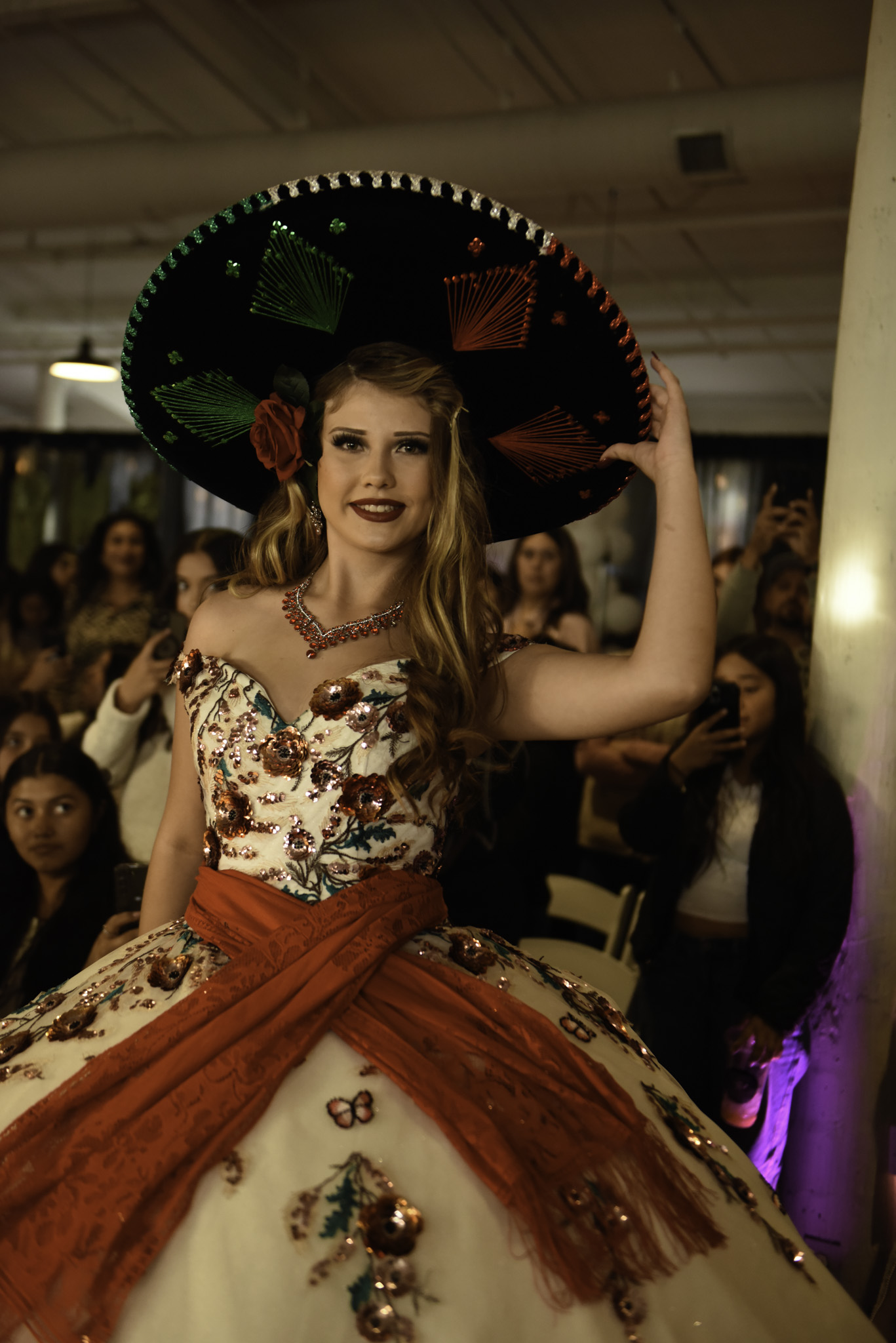 Young woman in traditional dress with a large sombrero, smiling at a festive event, surrounded by people.