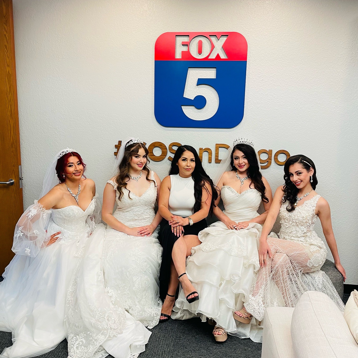 Five women in elegant dresses sit together in front of a FOX 5 sign, smiling and posing for the photo.