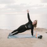 Woman exercising outdoors on a mat near water, raising one arm, with water and cloudy sky in background.