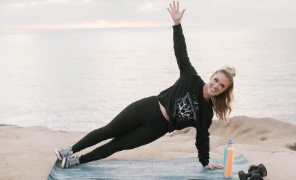 Woman exercising outdoors on a mat near water, raising one arm, with water and cloudy sky in background.