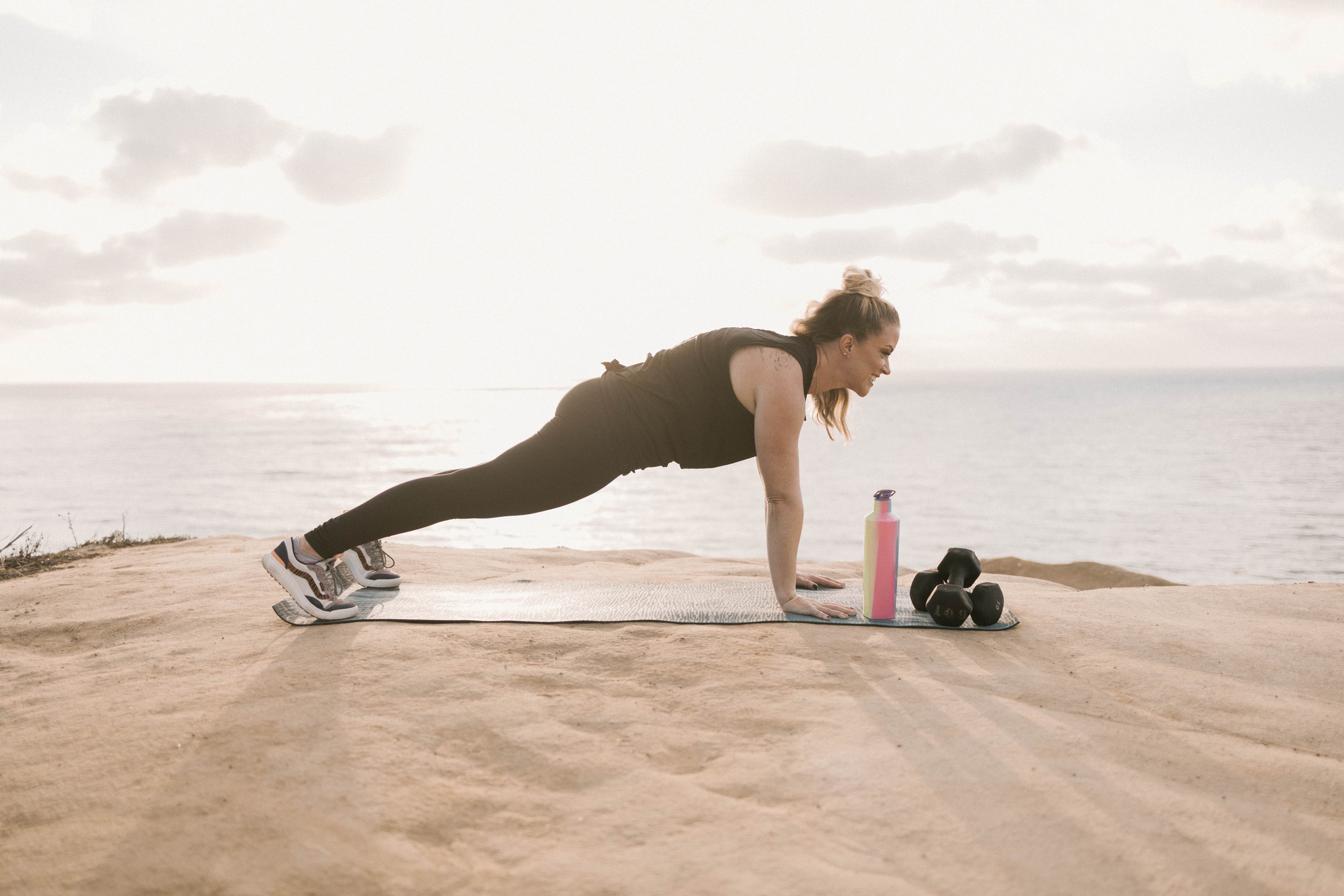 Person doing push-up on a mat outdoors near water, with water bottles and dumbbells nearby, during cloudy weather.