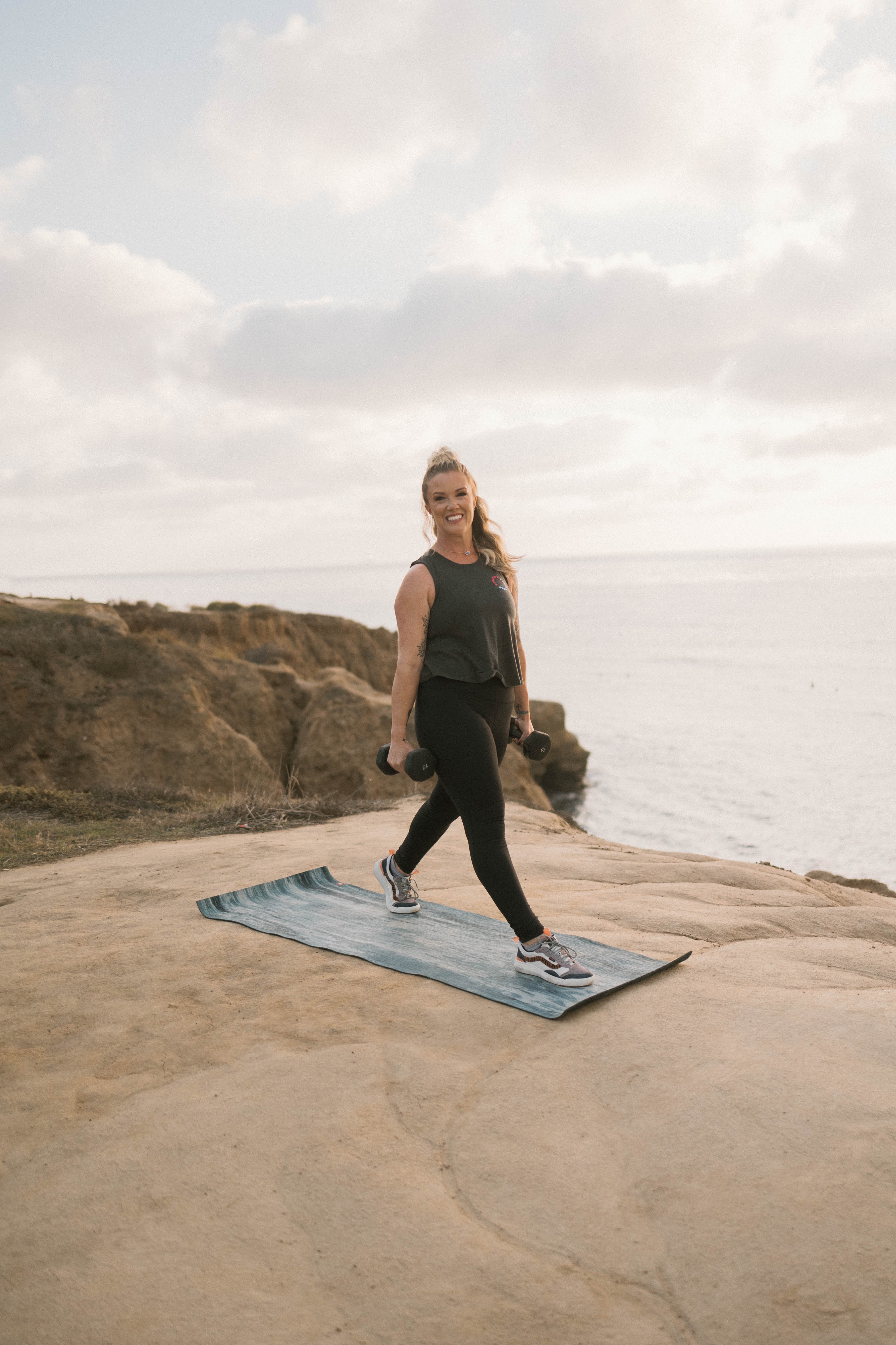 Woman walking on a mat outdoors near cliffs and ocean, cloudy sky, casual clothing, holding a dumbbell, smiling.