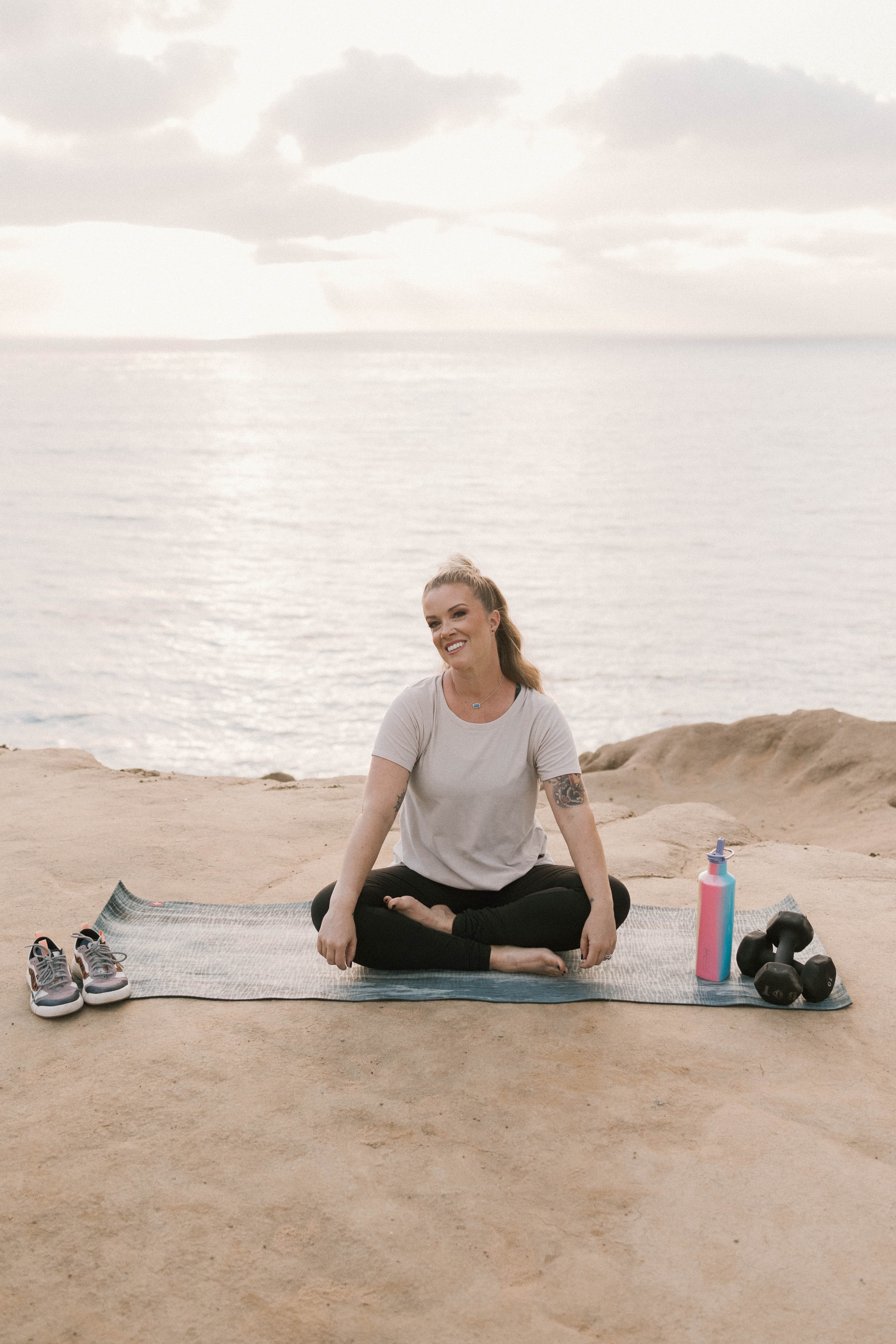 Woman practicing yoga on a beach with water and sky in background, sitting cross-legged on a mat, smiling.